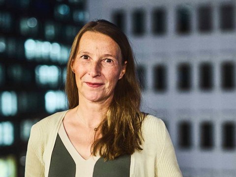 "Porträt einer Frau mit mittellangen, braunen Haaren und Sommersprossen. Sie trägt ein dunkelgrünes Oberteil unter einer hellen, gerippten Strickjacke und blickt lächelnd in die Kamera. Im Hintergrund ist ein unscharfes Hochhaus mit beleuchteten Fenstern und Fassadenelementen bei Nacht zu sehen."