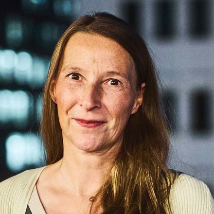 "Porträt einer Frau mit mittellangen, braunen Haaren und Sommersprossen. Sie trägt ein dunkelgrünes Oberteil unter einer hellen, gerippten Strickjacke und blickt lächelnd in die Kamera. Im Hintergrund ist ein unscharfes Hochhaus mit beleuchteten Fenstern und Fassadenelementen bei Nacht zu sehen."
