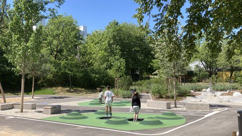 Zwei Personen stehen auf einem Spielfeld mit grünen und schwarzen Mustern umgeben von Bäumen und Bänken unter blauem Himmel