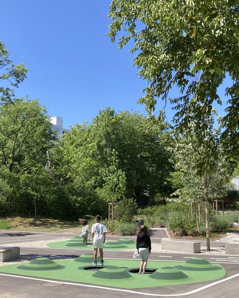 Zwei Personen stehen auf einem Spielfeld mit grünen und schwarzen Mustern umgeben von Bäumen und Bänken unter blauem Himmel