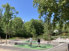 Zwei Personen stehen auf einem Spielfeld mit grünen und schwarzen Mustern umgeben von Bäumen und Bänken unter blauem Himmel