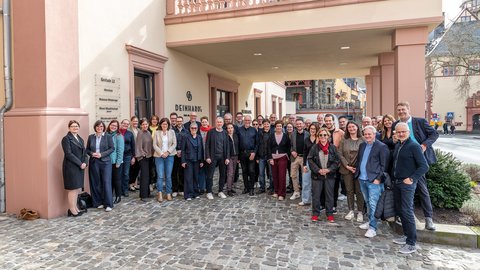 Gruppe von etwa 50 Personen steht vor einem historischen Gebäude mit Bogen und Pflastersteinboden