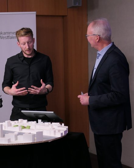 Zwei Personen stehen an einem runden Tisch mit weißem Architekturmodell; eine Person gestikuliert, hinter ihnen ein Roll‑up „Architektenkammer Nordrhein‑Westfalen“.