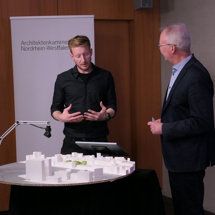 Zwei Personen stehen an einem runden Tisch mit weißem Architekturmodell; eine Person gestikuliert, hinter ihnen ein Roll‑up „Architektenkammer Nordrhein‑Westfalen“.