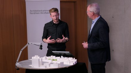 Zwei Personen stehen an einem runden Tisch mit weißem Architekturmodell; eine Person gestikuliert, hinter ihnen ein Roll‑up „Architektenkammer Nordrhein‑Westfalen“.