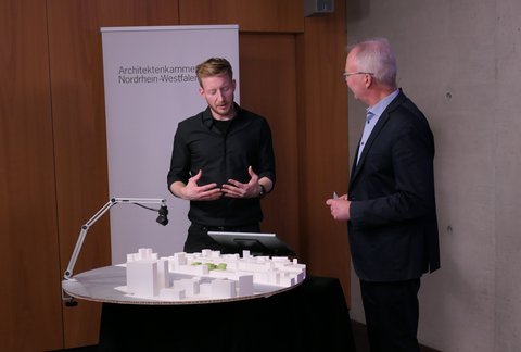 Zwei Personen stehen an einem runden Tisch mit weißem Architekturmodell; eine Person gestikuliert, hinter ihnen ein Roll‑up „Architektenkammer Nordrhein‑Westfalen“.