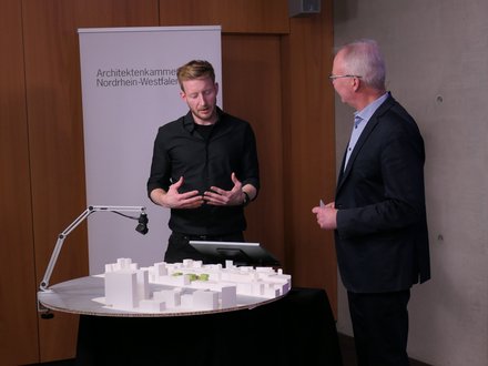 Zwei Personen stehen an einem runden Tisch mit weißem Architekturmodell; eine Person gestikuliert, hinter ihnen ein Roll‑up „Architektenkammer Nordrhein‑Westfalen“.