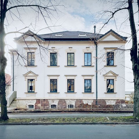 "Frontansicht eines weißen klassizistischen Gebäudes mit zwei Geschossen. Fünf große rechteckige Fenster mit Gewänden sind in zwei Reihen angeordnet. Das Dach ist dunkelblau mit Dachfenstern. Ein Sockelgeschoss aus rötlichem Stein und eine Treppe mit Metallgeländer sind zu sehen. Karge Bäume rahmen das Gebäude ein."
