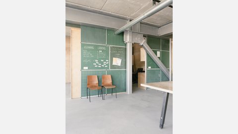 Innenraum mit grünen Tafel-Paneelen voller Notizen, zwei Holzstühle an der Wand; sichtbare Stahlträger.