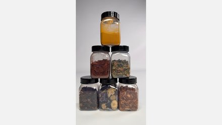 Sechs Kunststoffdosen als Pyramide: oben gelbes Pulver, darunter rote Späne und getrocknete Blätter, unten dunkle Rinden‑ und Pflanzenteile.