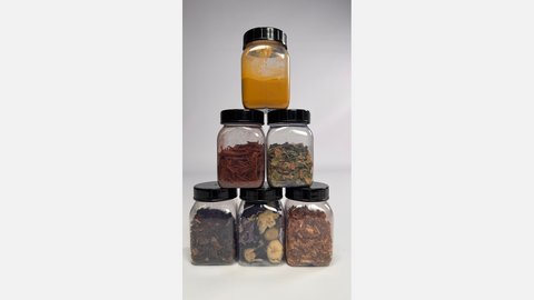 Sechs Kunststoffdosen als Pyramide: oben gelbes Pulver, darunter rote Späne und getrocknete Blätter, unten dunkle Rinden‑ und Pflanzenteile.