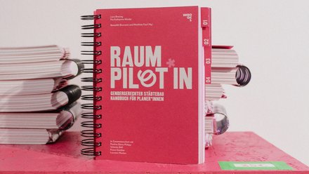 Das Bild zeigt ein rotes, spiralgebundenes Handbuch mit dem Titel „Raumpilotin – Gendergerechter Städtebau. Handbuch für Planerinnen“. Das Buch steht auf einer Fläche, daneben sind mehrere Exemplare gestapelt. Zweck des Bildes ist es, eine Publikation sichtbar zu machen, die Fachwissen und praktische Werkzeuge für gendergerechte Stadtplanung vermittelt.
