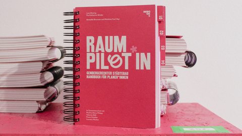 Das Bild zeigt ein rotes, spiralgebundenes Handbuch mit dem Titel „Raumpilotin – Gendergerechter Städtebau. Handbuch für Planerinnen“. Das Buch steht auf einer Fläche, daneben sind mehrere Exemplare gestapelt. Zweck des Bildes ist es, eine Publikation sichtbar zu machen, die Fachwissen und praktische Werkzeuge für gendergerechte Stadtplanung vermittelt.