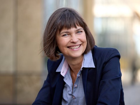 Halbnahes Porträt einer lächelnden erwachsenen Person mit kurzem braunem Haar, im dunkelblauen Blazer und hellblauen Hemd, draußen vor unscharfem, urbanem Hintergrund, Blick in die Kamera.