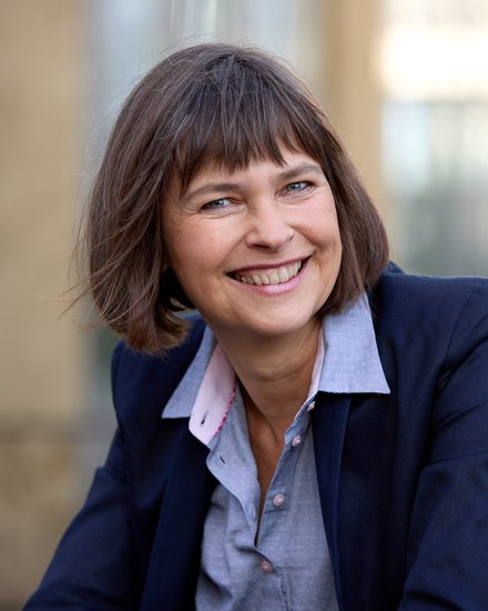 Halbnahes Porträt einer lächelnden erwachsenen Person mit kurzem braunem Haar, im dunkelblauen Blazer und hellblauen Hemd, draußen vor unscharfem, urbanem Hintergrund, Blick in die Kamera.