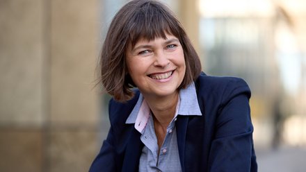 Halbnahes Porträt einer lächelnden erwachsenen Person mit kurzem braunem Haar, im dunkelblauen Blazer und hellblauen Hemd, draußen vor unscharfem, urbanem Hintergrund, Blick in die Kamera.