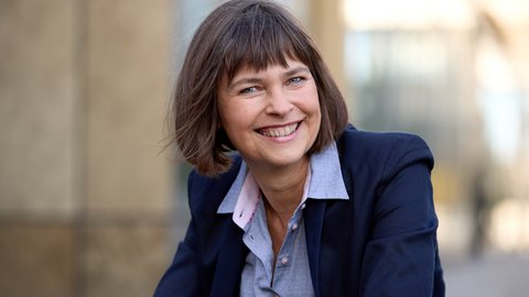 Halbnahes Porträt einer lächelnden erwachsenen Person mit kurzem braunem Haar, im dunkelblauen Blazer und hellblauen Hemd, draußen vor unscharfem, urbanem Hintergrund, Blick in die Kamera.