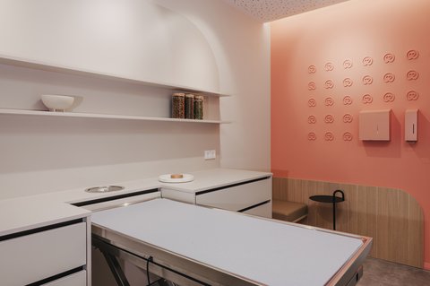 „Ein modernes Behandlungszimmer mit einem Behandlungstisch aus Edelstahl auf der linken Seite und einer markanten lachsfarbenen Wand mit Herz-Logos auf der rechten Seite.“