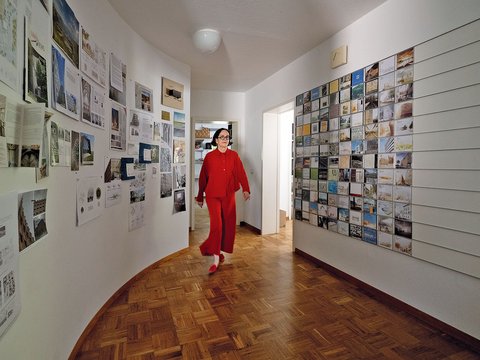 "Eine Frau in rotem Anzug geht durch einen langen, hellen Ausstellungsflur mit Holzparkett. Die Wände sind beidseitig mit zahlreichen architektonischen Plänen, Fotografien und Entwurfszeichnungen in schwarzen und beigen Rahmen tapeziert. Die linke Wand ist hellgrün gestrichen, die rechte Wand weiß. Ein weißer Deckenlüfter ist sichtbar. Die Ausstellung präsentiert ein umfassendes Spektrum von Architekturprojekten und Designkonzepten."