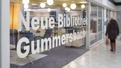 "Eingangsbereich einer modernen Bibliothek. Große Glasschiebetüren mit weißem Text "Neue Bibliothek Gummersbach". Dahinter Innenraum mit gelber Deckenakzent und weißen Kugelleuchten. Links ein gelber Sessel. Rechts durch die Glasfront sichtbar: Regale und Möbel. Dunkler Bodenbelag vor dem Eingang. Eine Person in dunkler Kleidung steht rechts neben der Tür."