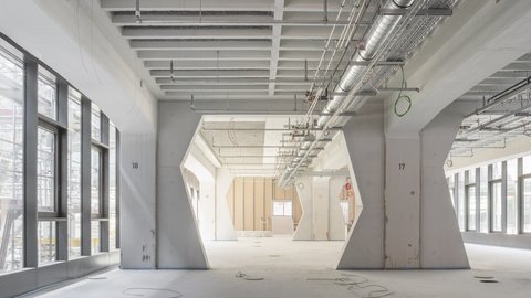 Großer Innenraum mit markanten Betonstützen  Ein weiter, heller Innenraum mit hohen Decken und regelmäßig angeordneten, massiven Betonstützen. Die Stützen haben eine besondere Form: Sie sind unten breit und verjüngen sich nach oben. An der Decke verlaufen sichtbare Lüftungsrohre, Kabeltrassen und technische Installationen. Links und rechts ziehen sich lange Fensterbänder entlang, die viel Tageslicht hereinlassen. Das Bild zeigt die industrielle Tragstruktur des Gebäudes. Die Offenheit und Sichtbarkeit der Technik verweisen auf Flexibilität und auf den Charakter eines Arbeits- oder Produktionsraums.