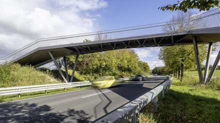 Moderne Fuß- und Radwegbrücke aus Stahl mit schlanken, geneigten Stützen überspannt eine zweispurige Straße; im Vordergrund fahren Autos mit Bewegungsunschärfe unter der Brücke hindurch, beidseitig gesäumt von grünen Bäumen und Leitplanken, blauer Himmel mit Wolken im Hintergrund.