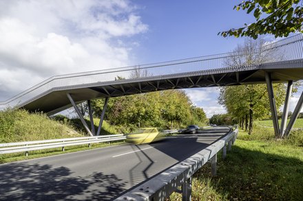 Moderne Fuß- und Radwegbrücke aus Stahl mit schlanken, geneigten Stützen überspannt eine zweispurige Straße; im Vordergrund fahren Autos mit Bewegungsunschärfe unter der Brücke hindurch, beidseitig gesäumt von grünen Bäumen und Leitplanken, blauer Himmel mit Wolken im Hintergrund.
