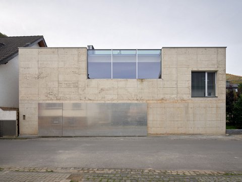 Frontalansicht eines minimalistischen Gebäudes mit Sichtbetonfassade, breitem Metalltor und Fensterfront im Obergeschoss.