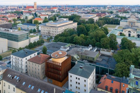 Luftaufnahme eines Stadtviertels mit mehreren Gebäuden, einem großen Park und einer weitläufigen Stadtlandschaft im Hintergrund