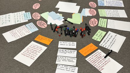 "Draufsicht auf einen grauen Teppichboden mit verteilten Arbeitmaterialien für einen Workshop oder Brainstorming-Prozess. In der Mitte steht ein Behälter mit schwarzen Stiften. Ringsum verteilt sind handgeschriebene und bedruckte Notizzettel in Weiß, Pastellgrün, Hellblau und Orange. Mehrere ovale rosa Klebepunkte mit Schrift markieren wichtige Punkte. Die Anordnung zeigt einen kreativen Organisationsprozess mit Ideen und Konzepten."  Bild 2: Team bei Wh