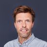 Ein farbiges Porträtfoto desselben Mannes wie in Bild 1 vor einem einheitlichen, dunklen graublauen Hintergrund. Der Mann hat kurze, bräunliche Haare, blaue Augen und einen Stoppelbart. Er trägt ein hellgraues oder graublaues Hemd mit Button-Down-Kragen. Er schaut direkt in die Kamera und hat einen ähnlichen Gesichtsausdruck wie in der Skizze.