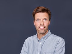 Ein farbiges Porträtfoto desselben Mannes wie in Bild 1 vor einem einheitlichen, dunklen graublauen Hintergrund. Der Mann hat kurze, bräunliche Haare, blaue Augen und einen Stoppelbart. Er trägt ein hellgraues oder graublaues Hemd mit Button-Down-Kragen. Er schaut direkt in die Kamera und hat einen ähnlichen Gesichtsausdruck wie in der Skizze.