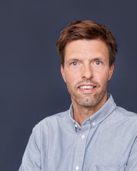 Ein farbiges Porträtfoto desselben Mannes wie in Bild 1 vor einem einheitlichen, dunklen graublauen Hintergrund. Der Mann hat kurze, bräunliche Haare, blaue Augen und einen Stoppelbart. Er trägt ein hellgraues oder graublaues Hemd mit Button-Down-Kragen. Er schaut direkt in die Kamera und hat einen ähnlichen Gesichtsausdruck wie in der Skizze.