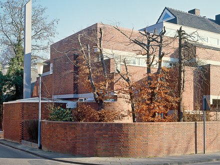 "Modernes Backsteingebäude mit Satteldach an einer Straßenecke. Rote Backsteinmauern, Gartenmauer aus Backstein, kahle Bäume. Ein dunkler Quader-Anbau mit verglaster Fassade auf der linken Seite. Klarer Himmel."