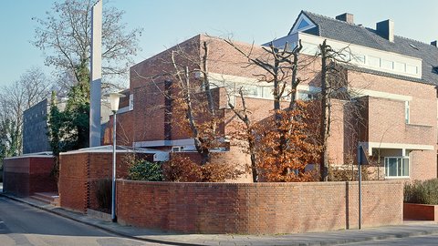 "Modernes Backsteingebäude mit Satteldach an einer Straßenecke. Rote Backsteinmauern, Gartenmauer aus Backstein, kahle Bäume. Ein dunkler Quader-Anbau mit verglaster Fassade auf der linken Seite. Klarer Himmel."