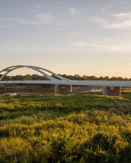 Eine weite Landschaftsaufnahme zeigt eine moderne Brücke mit elegantem Bogen über einen Fluss und angrenzende Auenlandschaft im warmen Licht, wodurch das Bild ein Infrastrukturprojekt als Teil von Landschaft und Raum darstellt und für die Verbindung von Ingenieurbau, Umwelt und regionaler Identität steht.