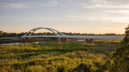 Eine weite Landschaftsaufnahme zeigt eine moderne Brücke mit elegantem Bogen über einen Fluss und angrenzende Auenlandschaft im warmen Licht, wodurch das Bild ein Infrastrukturprojekt als Teil von Landschaft und Raum darstellt und für die Verbindung von Ingenieurbau, Umwelt und regionaler Identität steht.