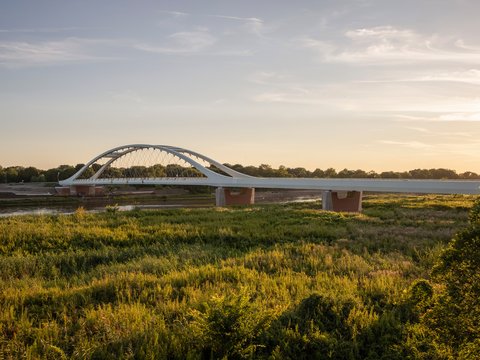 Eine weite Landschaftsaufnahme zeigt eine moderne Brücke mit elegantem Bogen über einen Fluss und angrenzende Auenlandschaft im warmen Licht, wodurch das Bild ein Infrastrukturprojekt als Teil von Landschaft und Raum darstellt und für die Verbindung von Ingenieurbau, Umwelt und regionaler Identität steht.