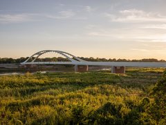 Eine weite Landschaftsaufnahme zeigt eine moderne Brücke mit elegantem Bogen über einen Fluss und angrenzende Auenlandschaft im warmen Licht, wodurch das Bild ein Infrastrukturprojekt als Teil von Landschaft und Raum darstellt und für die Verbindung von Ingenieurbau, Umwelt und regionaler Identität steht.