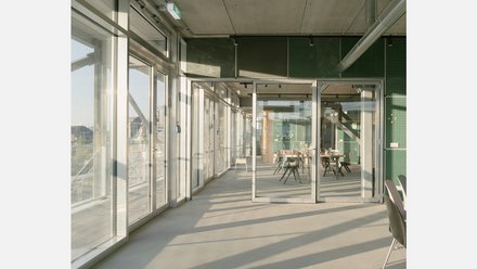 Flur mit bodentiefen Glasschiebetüren, grünen Wandpaneelen und sichtbaren Lüftungsrohren; hinten Besprechungsraum mit Tisch und Stühlen.