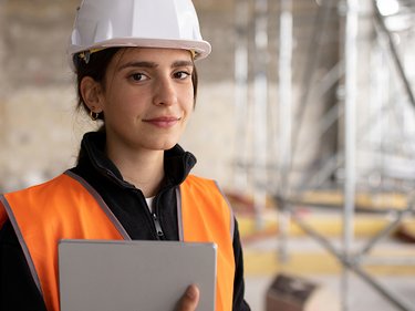 Person mit weißem Schutzhelm und orangefarbener Warnweste hält ein Tablet in einer Baustellenumgebung mit Gerüst im Hintergrund.