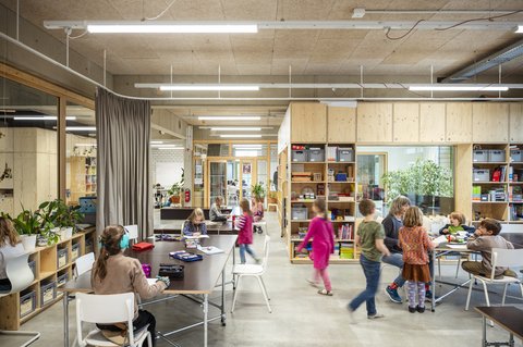 Heller Lernraum mit Holzeinbauten und offenen Regalen. Kinder sitzen an Tischen und malen, lesen oder arbeiten gemeinsam.