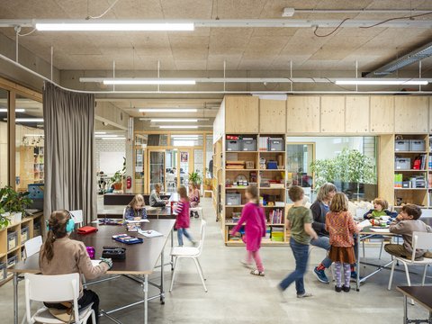 Heller Lernraum mit Holzeinbauten und offenen Regalen. Kinder sitzen an Tischen und malen, lesen oder arbeiten gemeinsam.