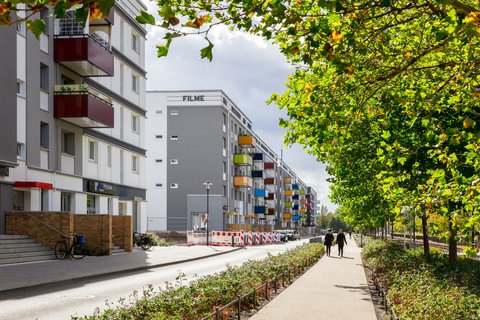 Sanierte Wohnanlage mit farbigen Balkonen entlang einer begrünten Straße; auf dem Gehweg gehen mehrere Menschen zwischen Bäumen.
