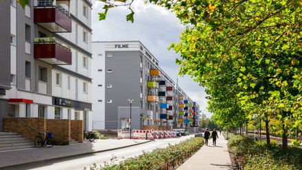 Sanierte Wohnanlage mit farbigen Balkonen entlang einer begrünten Straße; auf dem Gehweg gehen mehrere Menschen zwischen Bäumen.