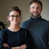 Zwei Personen stehen in einem Innenraum und lächeln in die Kamera; eine trägt Brille, beide in dunkler Kleidung.