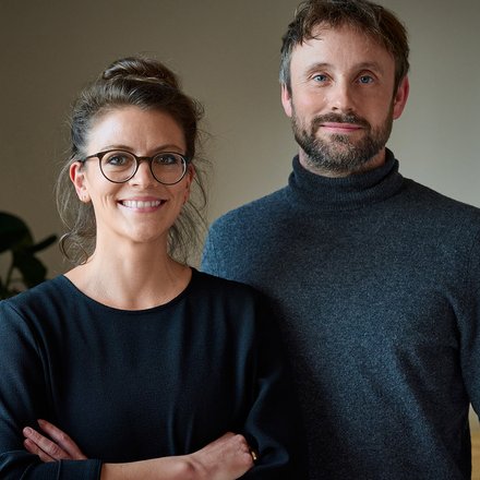 Zwei Personen stehen in einem Innenraum und lächeln in die Kamera; eine trägt Brille, beide in dunkler Kleidung.