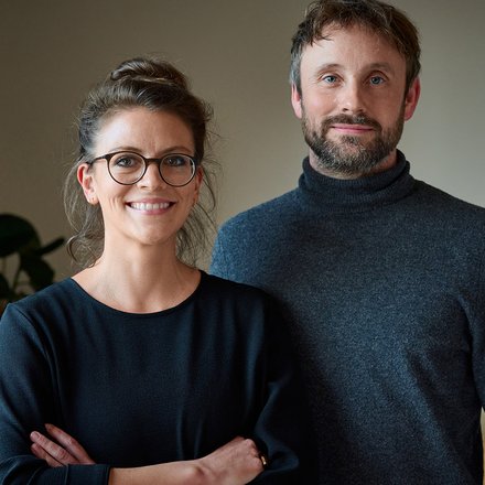Zwei Personen stehen in einem Innenraum und lächeln in die Kamera; eine trägt Brille, beide in dunkler Kleidung.