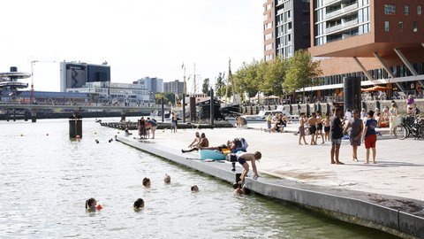Menschen schwimmen und entspannen an einer sonnigen Uferpromenade mit modernen Gebäuden, Booten und lebhafter Hafenatmosphäre im Hintergrund.