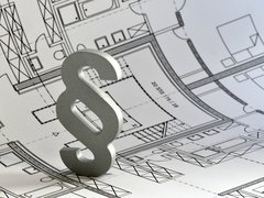 Ein silbernes Paragrafenzeichen steht auf einem ausgebreiteten architektonischen Grundrissplan